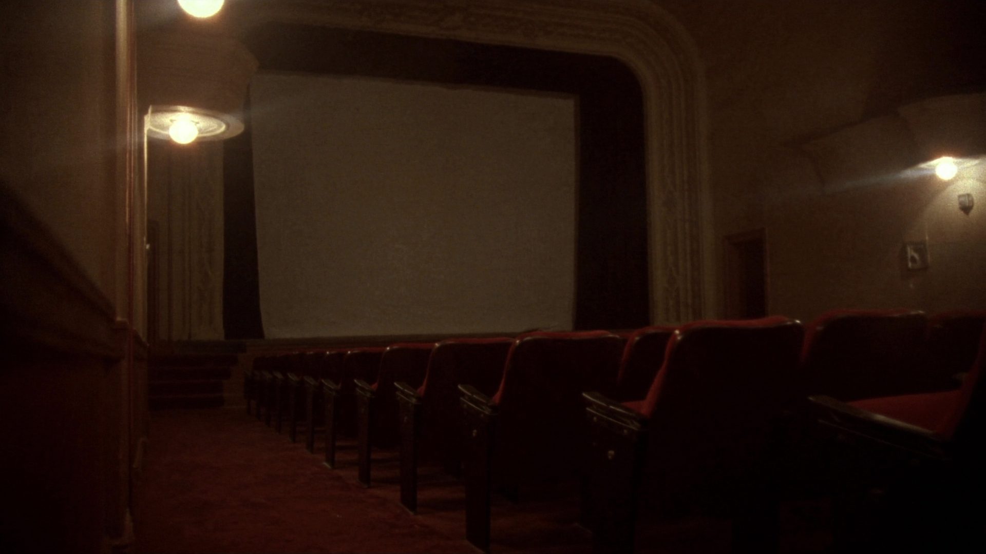 The interior of a restored historic single-screen cinema seen from the back of the house: rows of red upholstered seats sloping down toward a blank white screen inside an ornate plaster arch, house lights at half, a side exit lamp glowing.